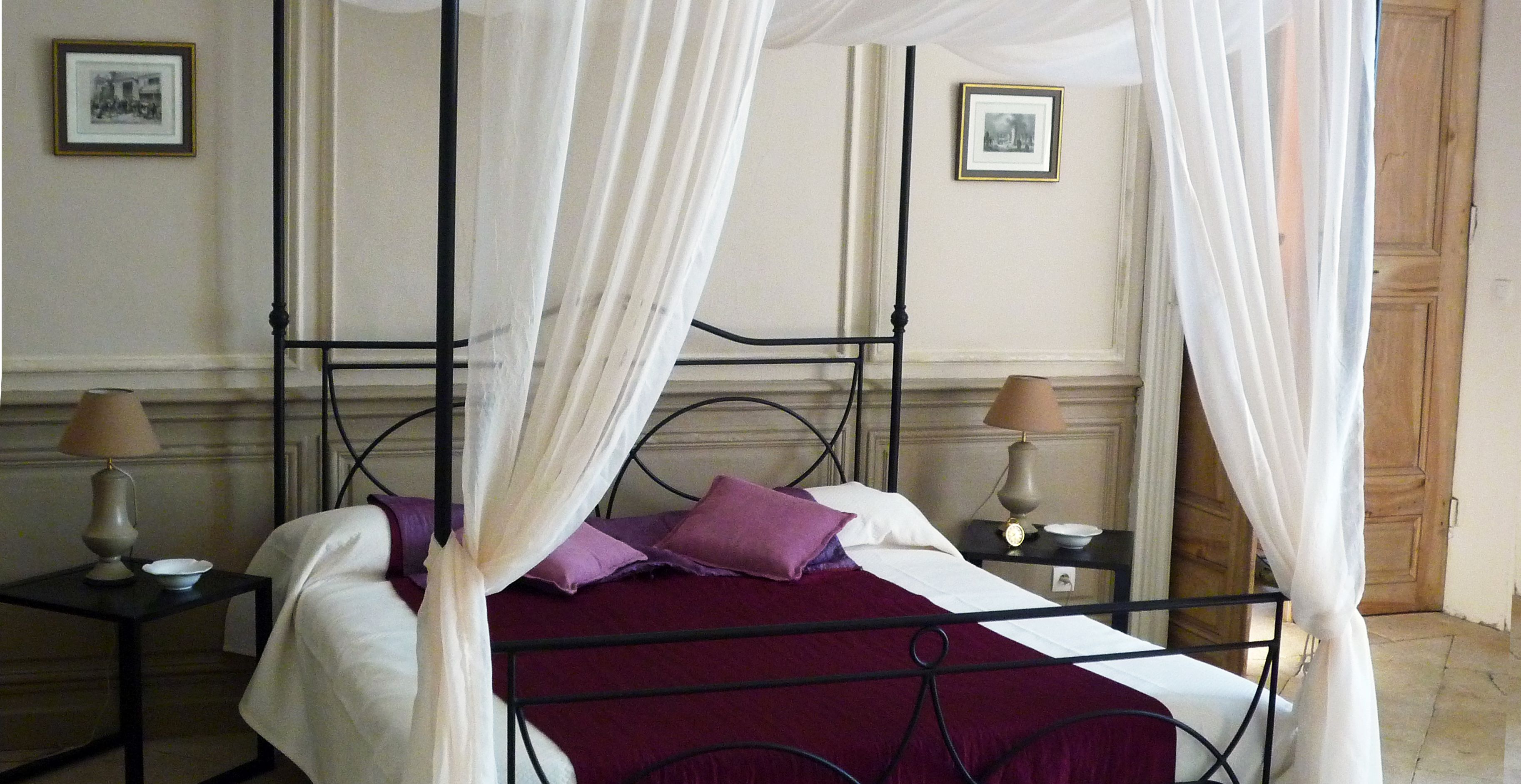 Una cama con dosel adornada con cortinas blancas, dos mesitas de noche con lámparas, y cuadros enmarcados en las paredes.