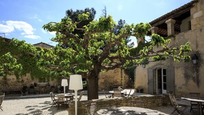 An outdoor patio with wooden tables and chairs, surrounded by stone walls and a large tree. - Domaine des Escaunes (Sernhac)