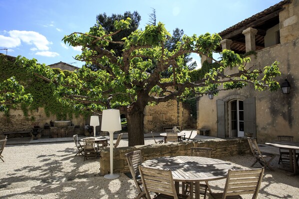 Un patio al aire libre con mesas y sillas de madera, rodeado de muros de piedra y un árbol grande.