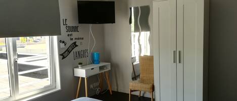A bedroom with a bed, a TV mounted on the wall, a wardrobe, a chair, and a small table with a lamp.