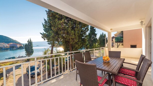 Un balcon agrémenté de meubles en osier, une table avec un panier de fruits et une vue sur la mer et les bâtiments.