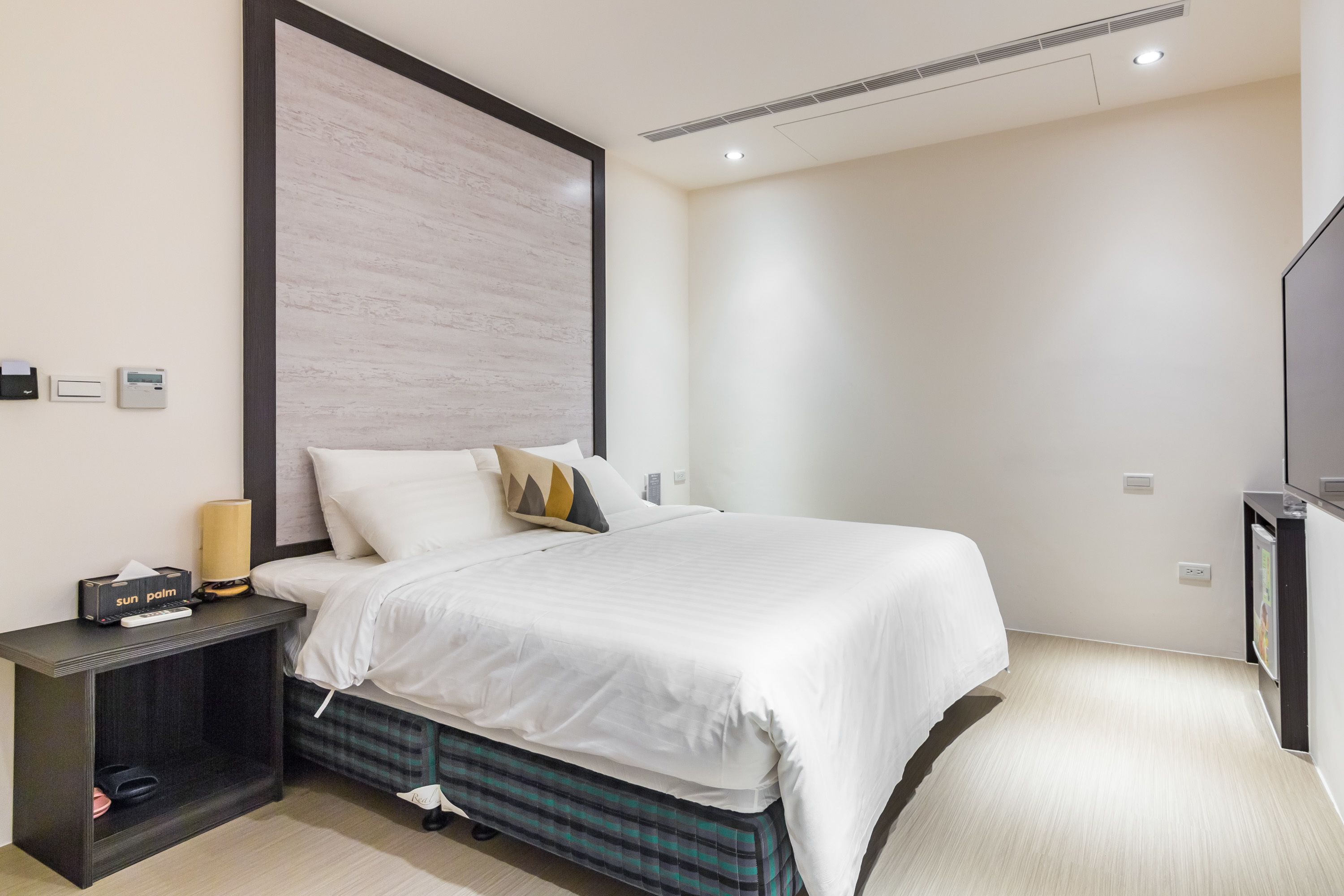 Una habitación de hotel moderna con una cama grande, una mesita de noche con lámpara y un televisor en la pared.