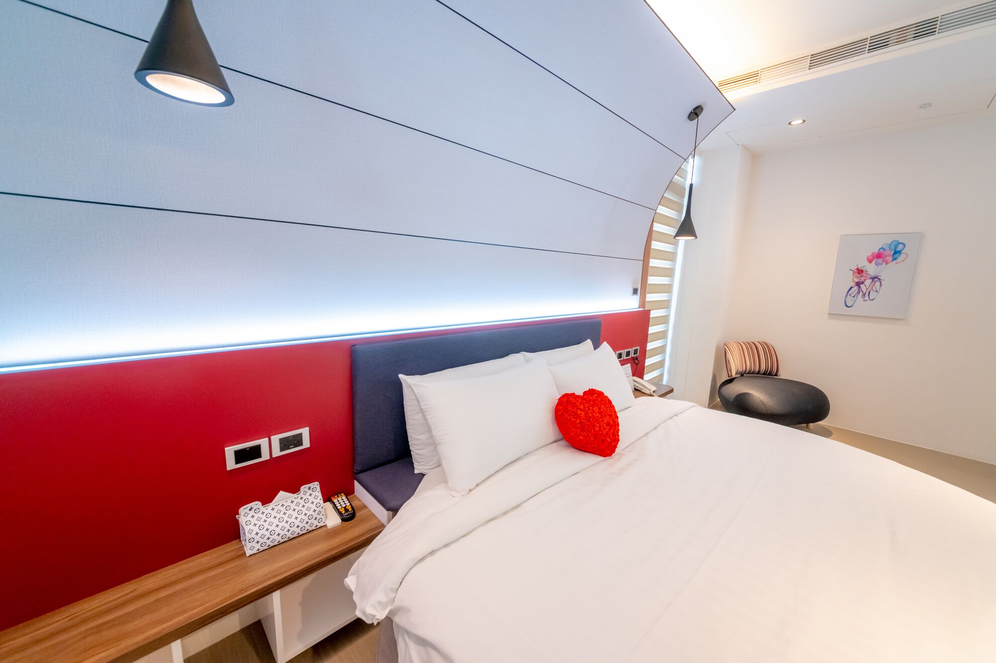 A modern hotel room with a red accent wall, a bed with white bedding, a heart-shaped pillow, a bedside table with a remote control, and a wall-mounted light fixture.
