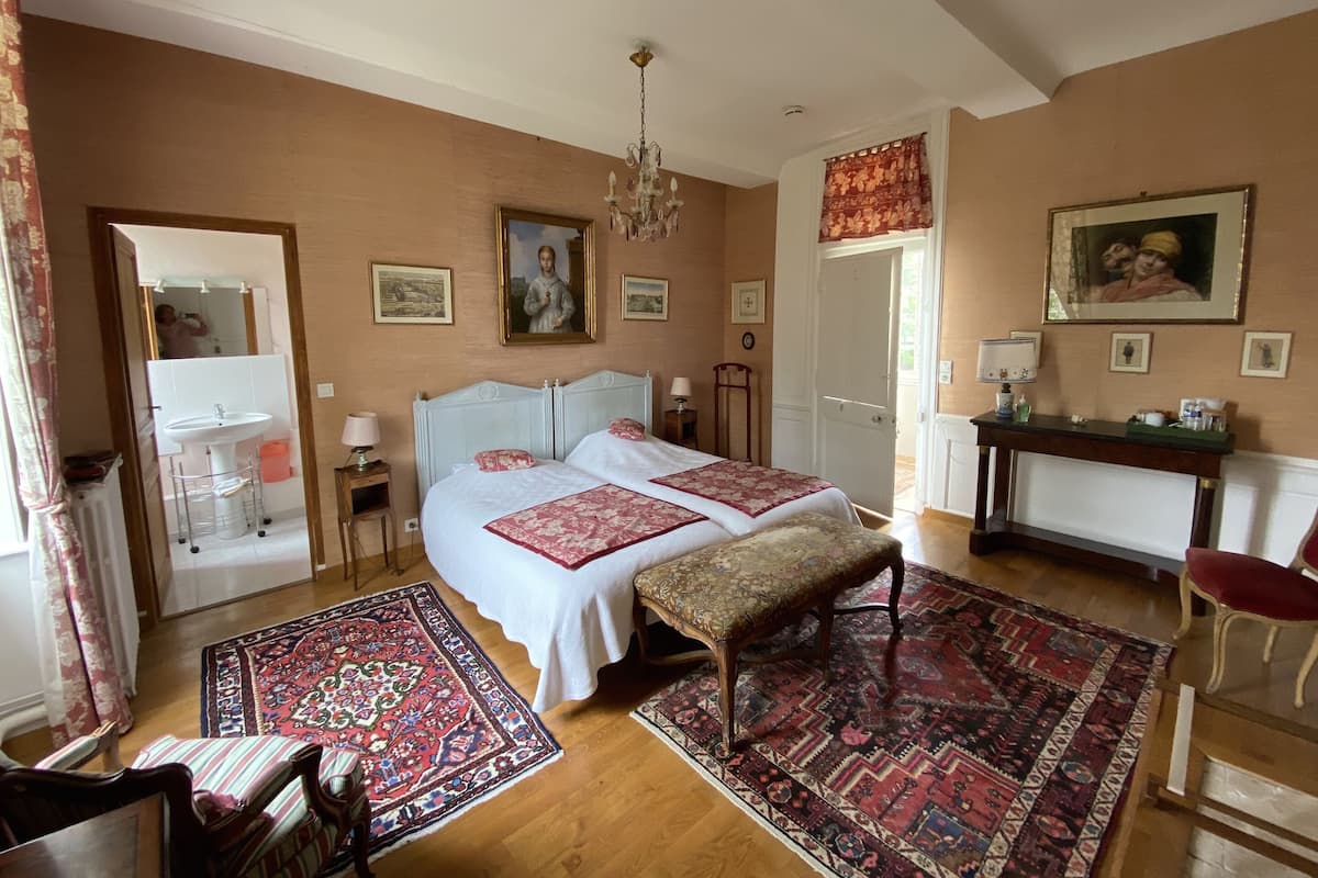 A bedroom with a large bed, a sitting area with a chair, a small table with a lamp, and a mirror.