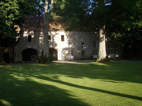 Un bâtiment historique en pierre, doté de nombreuses fenêtres et d’une entrée centrale, entouré d’une pelouse bien entretenue et d’arbres.