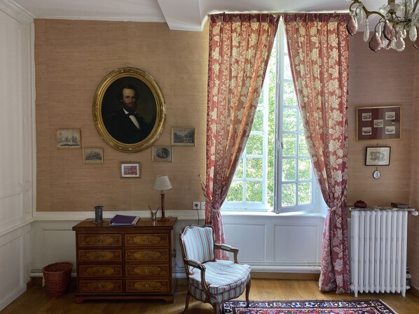 Quarto com um retrato grande, penteadeira de madeira, uma cadeira e uma janela com cortinas florais.