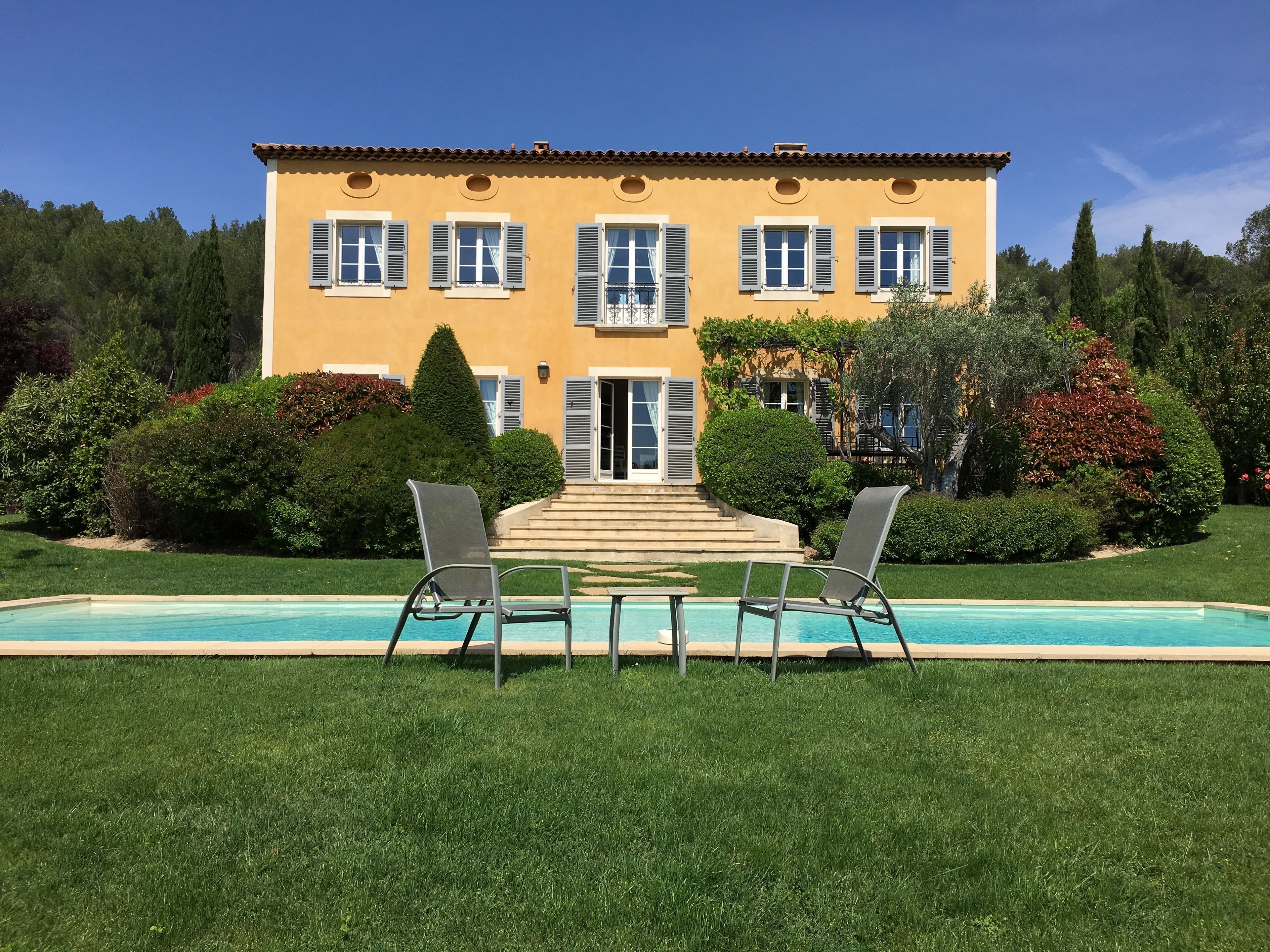 A yellow building with a swimming pool, two chairs, and a staircase leading to the entrance.