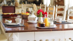 A breakfast setup with a coffee maker, teapot, cups, a fruit bowl, and a vase with red flowers on a wooden table.