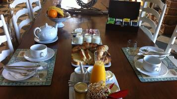 Une table en bois dressée pour le petit-déjeuner, avec une variété d’aliments et de boissons, entourée de chaises blanches.