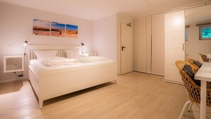 A modern bedroom with a white bed, bedside tables, a desk, and a chair. There is a wardrobe and a door leading to another room.