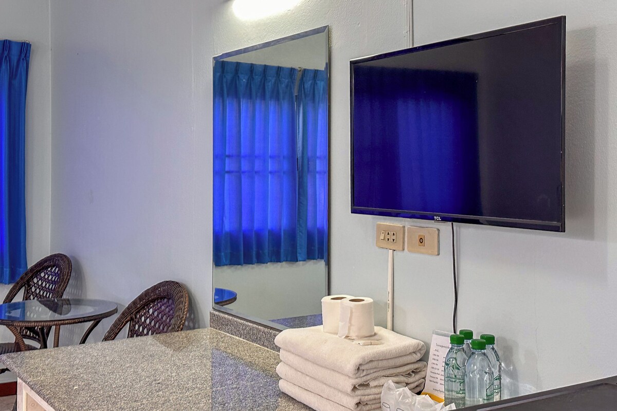 A hotel room with a flat-screen TV, a mirror, a small table with chairs, a stack of towels, and toiletries.