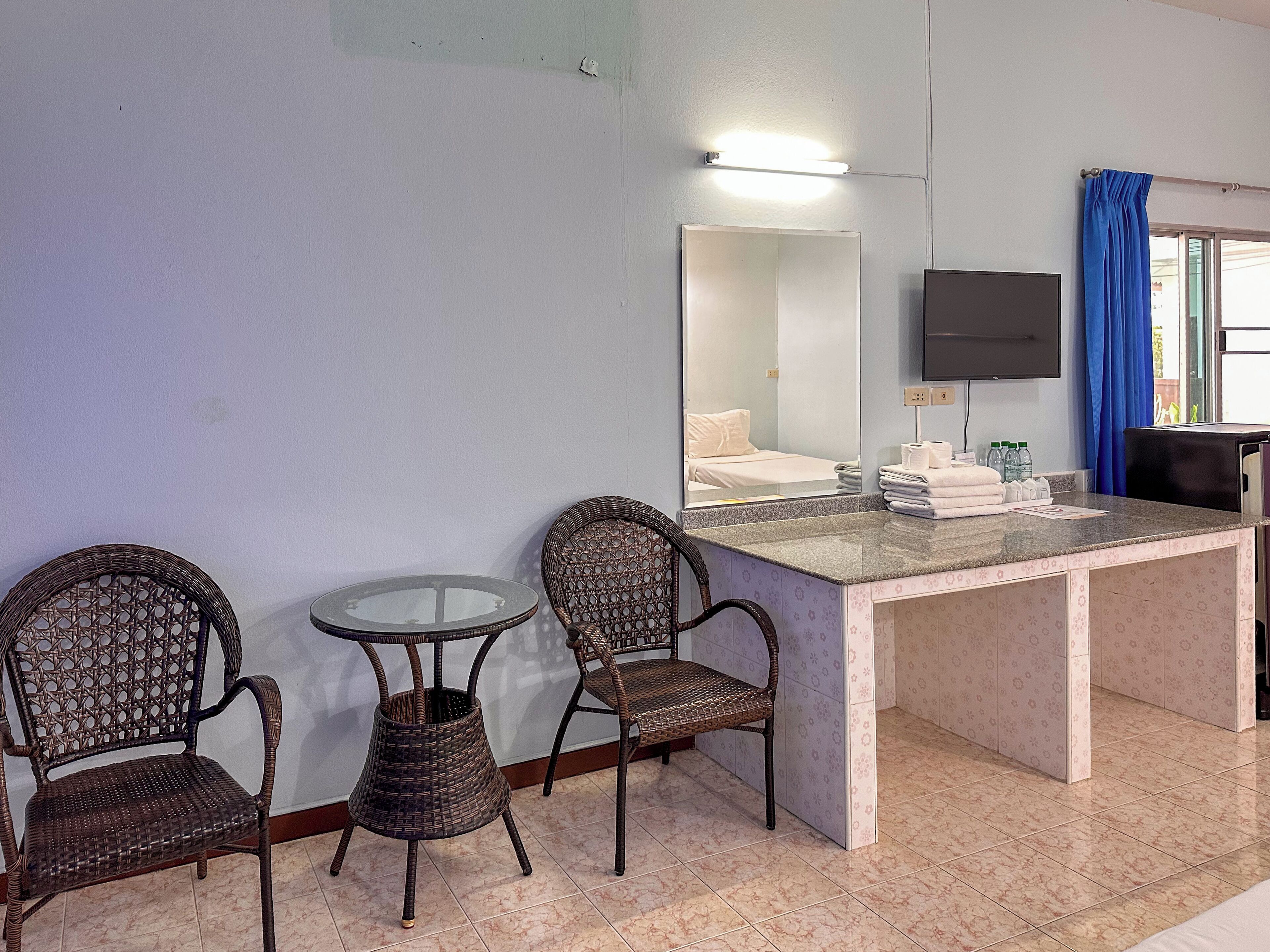 A hotel room with a bed, a small table, two wicker chairs, a mirror, a flat-screen TV, and a counter with towels and toiletries.