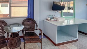 Hotelkamer met rieten stoelen, een glazen tafel, een bureau met toiletartikelen, een televisie en een raam met uitzicht op gebouwen.