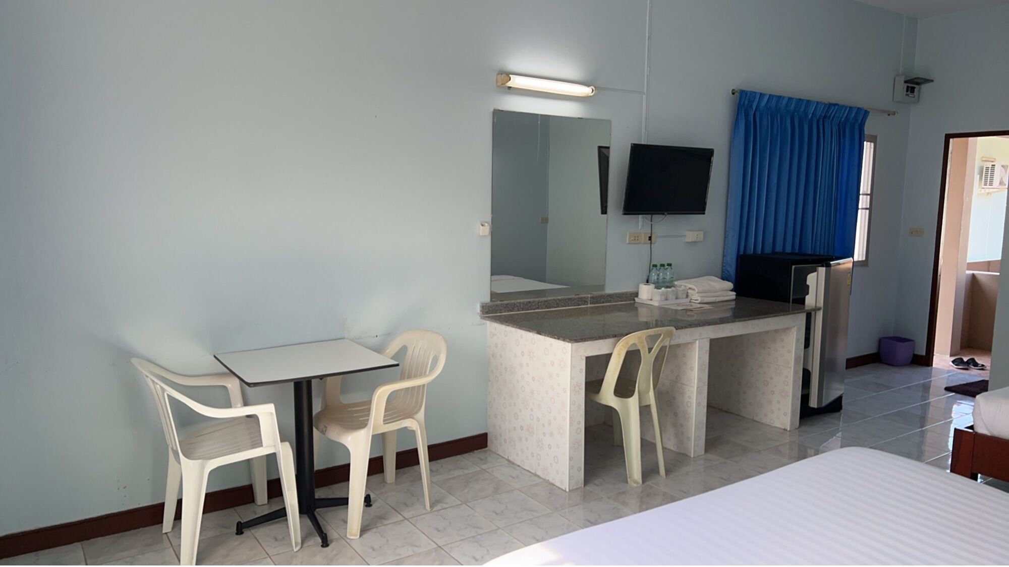 A hotel room with a bed, a desk, two chairs, a mirror, a TV, and a window with blue curtains.
