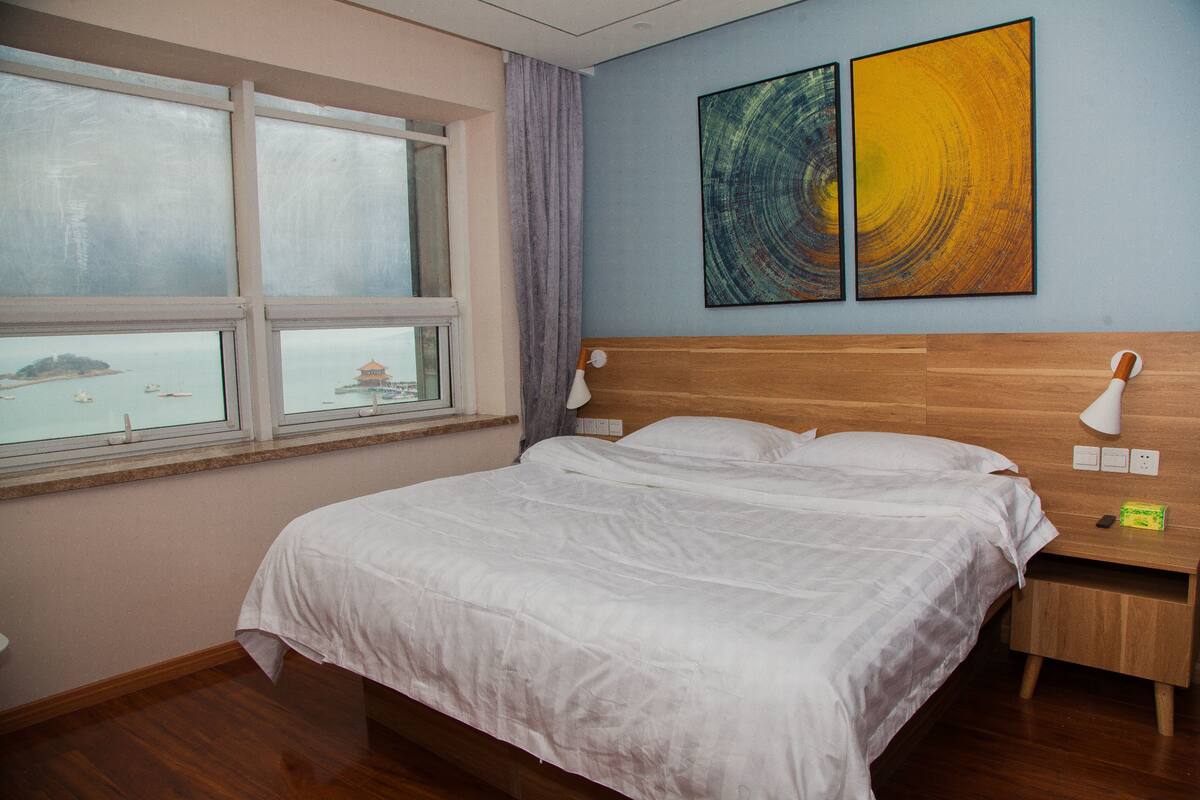 A bedroom with a large bed, a nightstand, a lamp, and a window with a view of a distant island.