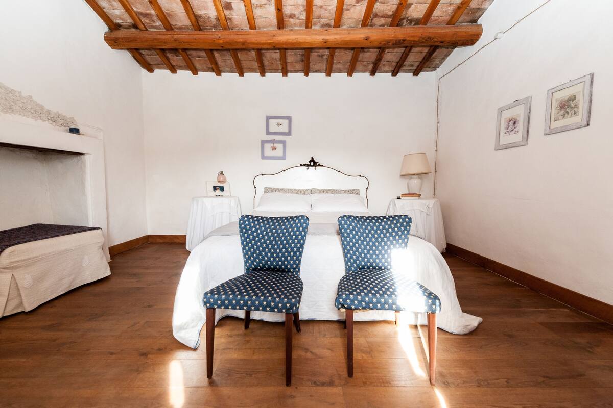 A bedroom with a wooden ceiling, a bed with a white headboard, two patterned chairs, a bedside table with a lamp, and a small picture on the wall.