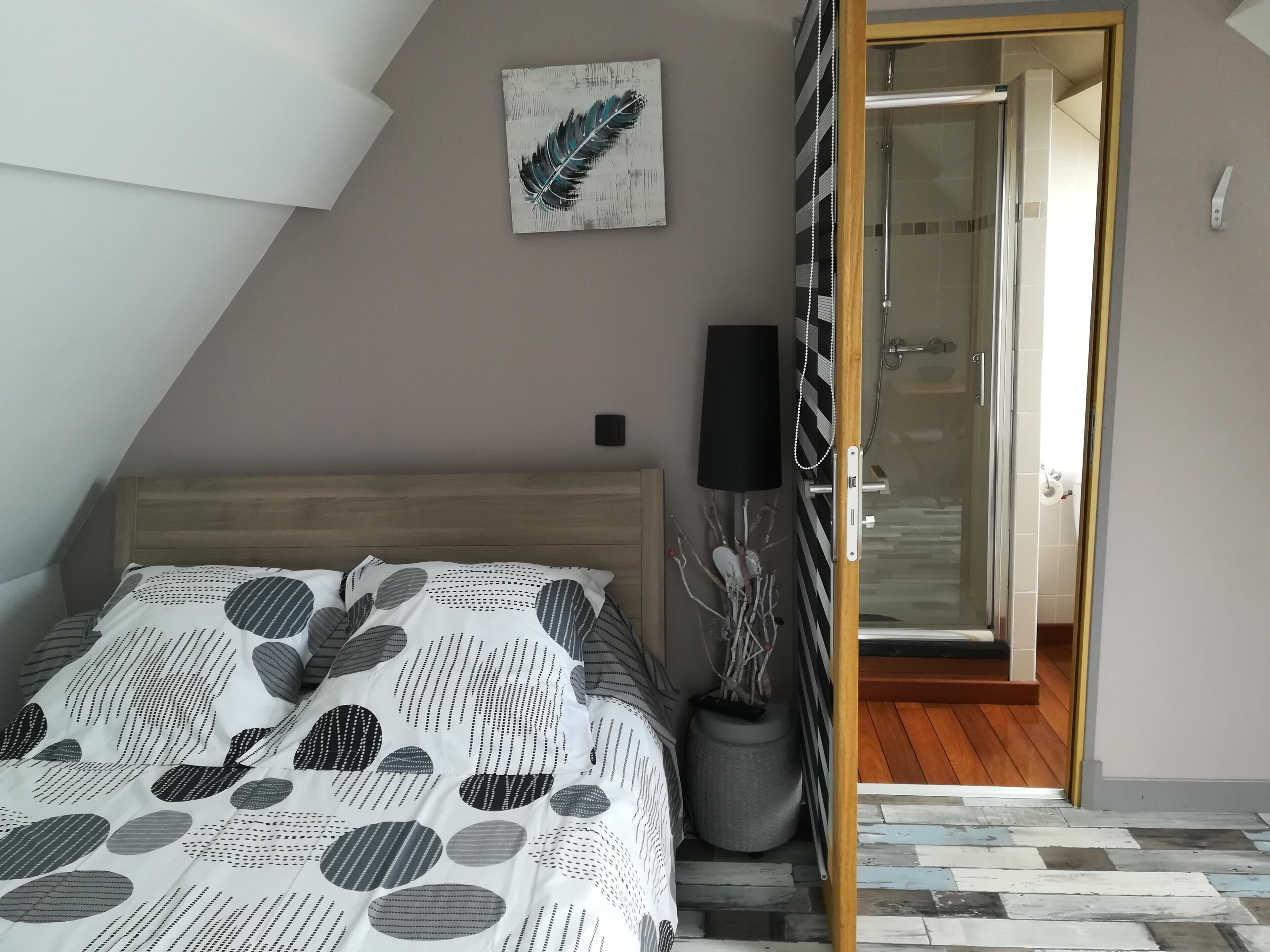 Een slaapkamer met een schuine plafond, een bed met een bedovertrek met patroon, een potplant, een speaker bevestigd aan de muur en een deur naar een badkamer.