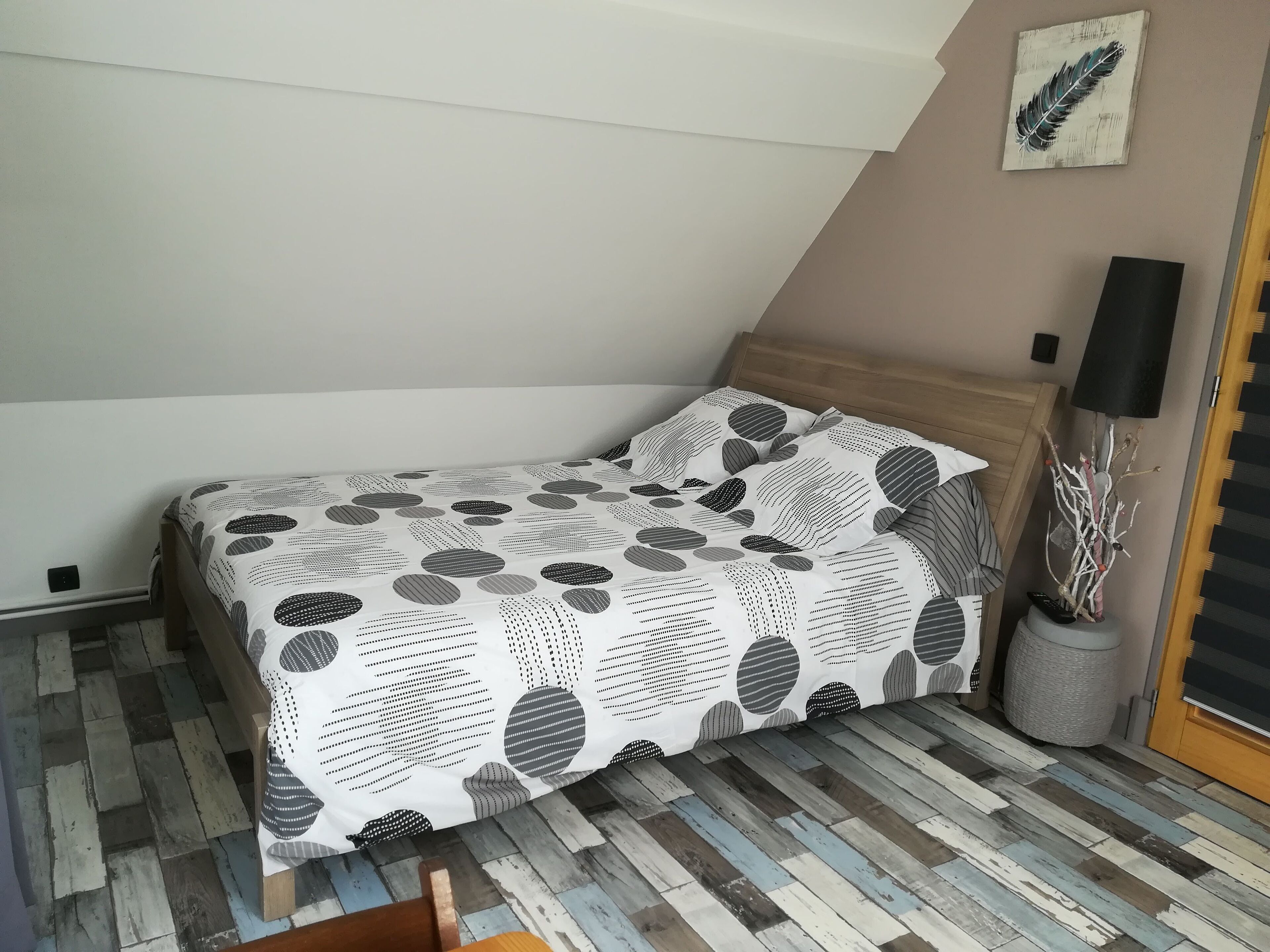 Ein Schlafzimmer mit Bett, Holz-Kopfteil, Wandlampe, Zimmerpflanze und einem gemusterten Bettbezug.