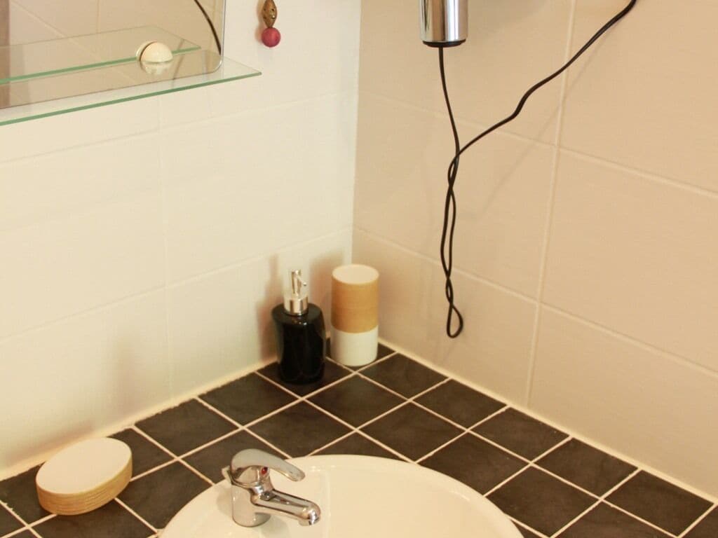 Un baño con un lavabo blanco, baldosas marrón oscuro, un estante de vidrio, un secador de pelo y un dispensador de papel higiénico.