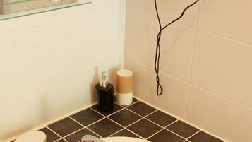 Un baño con un lavabo blanco, baldosas marrón oscuro, un estante de vidrio, un secador de pelo y un dispensador de papel higiénico.