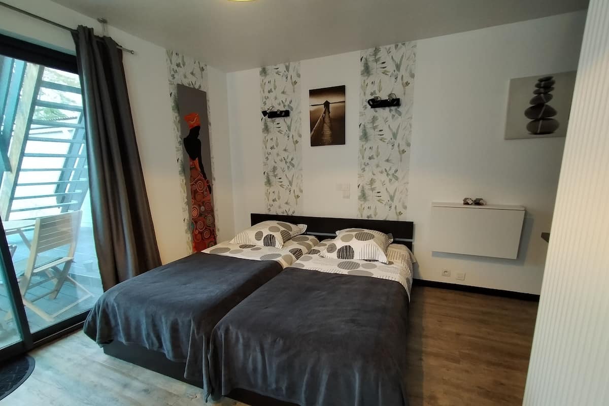A bedroom with two beds, a window with curtains, a wall-mounted radiator, and a small shelf with decorative items.