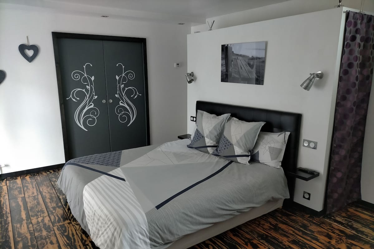 A modern bedroom with a large bed, a wardrobe with decorative doors, a small bedside table, and a wall-mounted light fixture.