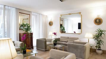 Uma sala de estar bem iluminada com um sofá, poltronas, uma mesa de centro e uma mesa com tampo de vidro. Há duas luminárias, um espelho e um vaso de planta.
