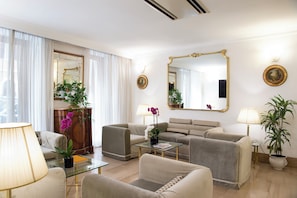 A well-lit living room with a sofa, armchairs, a coffee table, and a glass-top table. There are two lamps, a mirror, and a potted plant. - Hotel Cecil (Rome)