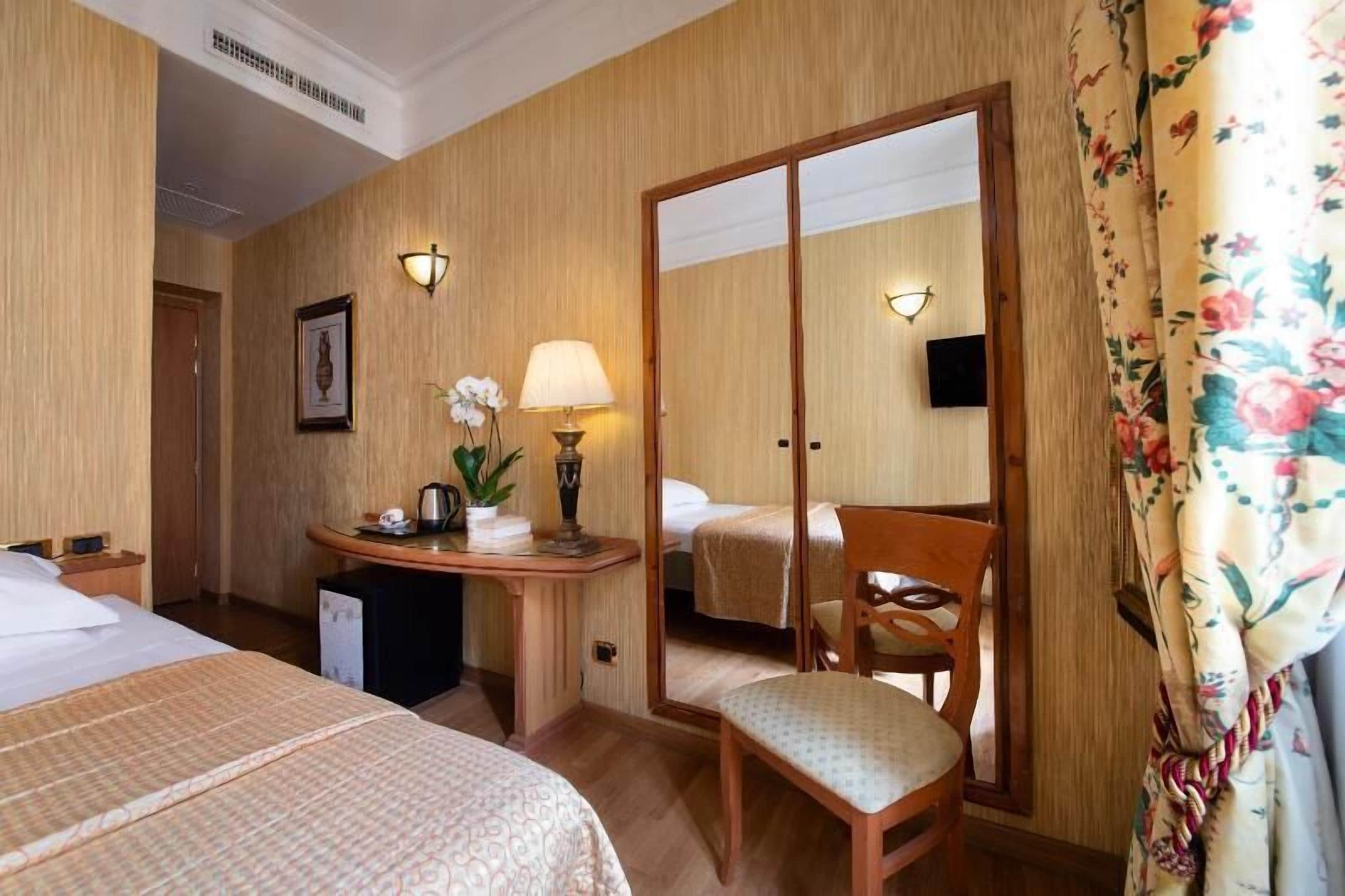 A hotel room with two beds, a desk with a lamp, a chair, and a mirror.