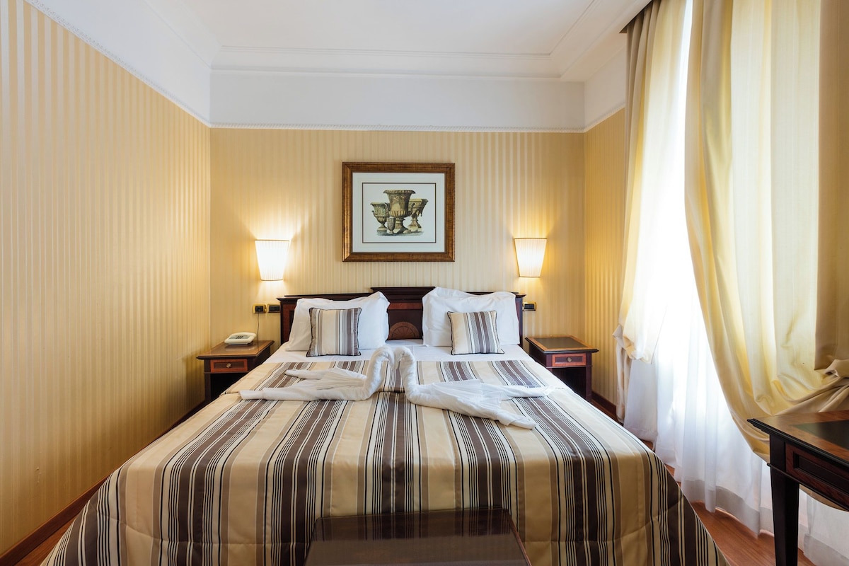 A hotel room with a large bed, two bedside tables, a nightstand, a framed picture on the wall, and a window with curtains.