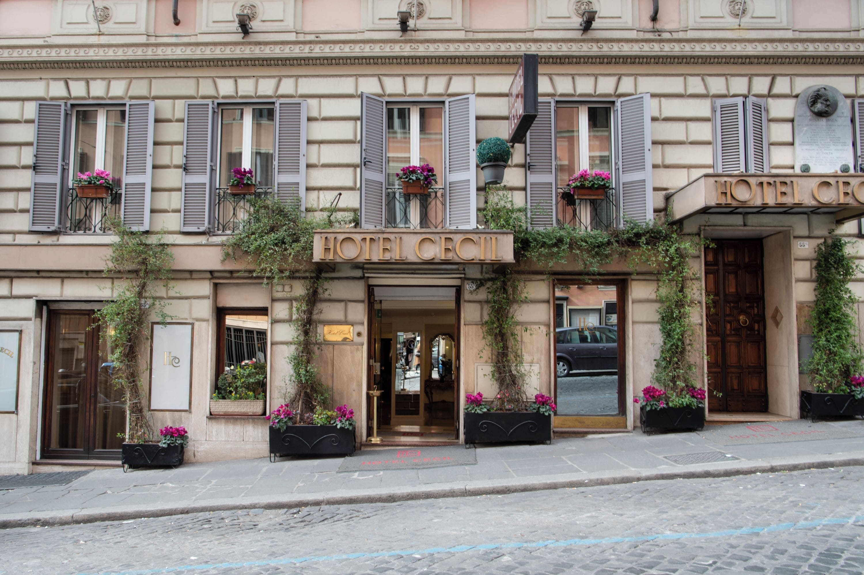 A hotel facade with a classic architectural style, featuring a prominent entrance, multiple windows with shutters, and decorative flower boxes.