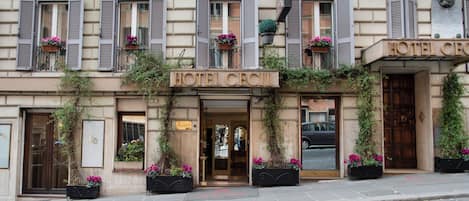 A hotel facade with a classic architectural style, featuring a prominent entrance, multiple windows with shutters, and decorative flower boxes.