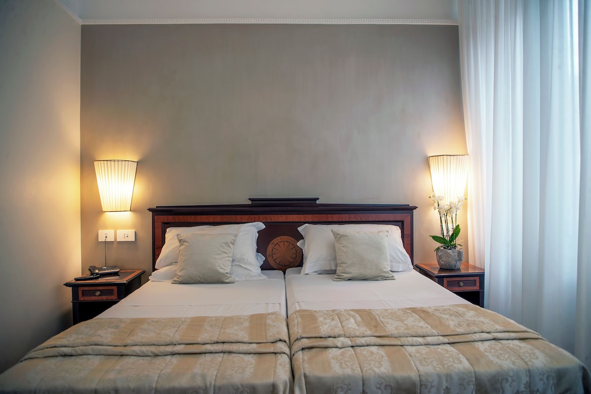 A double bed with a wooden headboard, two bedside tables with lamps, and a potted plant on the right side.