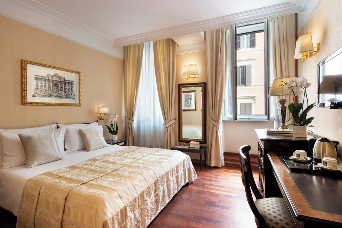 A hotel room with a large bed, a desk with a chair, a mirror, and a window with curtains.