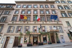 Fachada de hotel con múltiples ventanales, una entrada central y banderas de Italia, Francia y la UE.