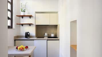 Una cocina compacta con armarios blancos, una cafetera, un frutero y una orquídea en maceta sobre una estantería.