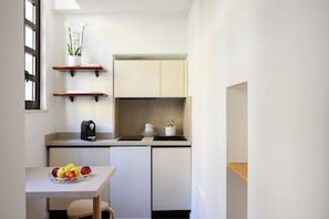 A compact kitchen with white cabinets, a coffee machine, a fruit bowl, and a potted orchid on a shelf. - Hotel Cecil (Rome)