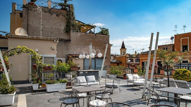An outdoor patio area with multiple tables and chairs, potted plants, and a building with a tower in the background. - Hotel Cecil (Rome)