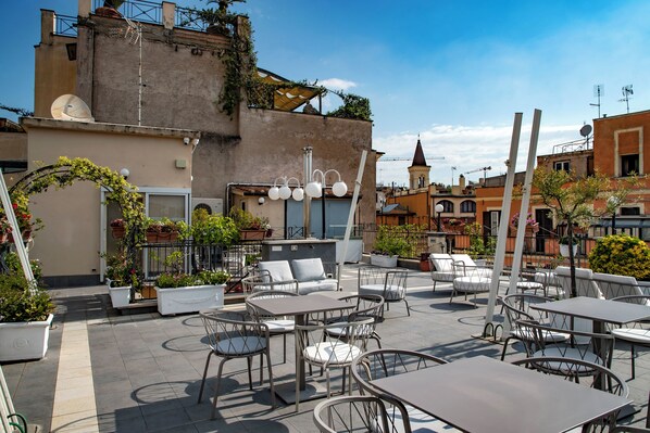 An outdoor patio area with multiple tables and chairs, potted plants, and a building with a tower in the background. - Hotel Cecil (Rome)
