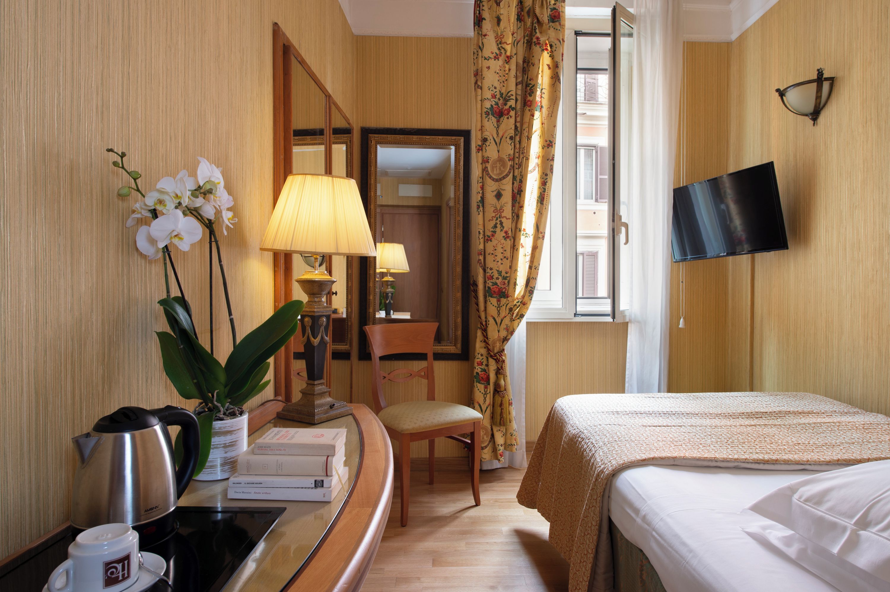 Una habitación de hotel con cama, escritorio con lámpara, silla, televisor y ventana con cortinas.