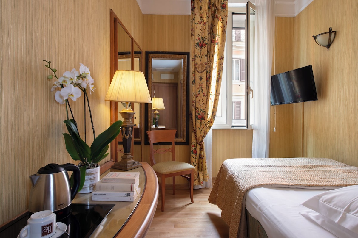 A hotel room with a bed, a desk with a lamp, a chair, a television, and a window with curtains.