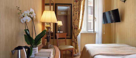 Una habitación de hotel con cama, escritorio con lámpara, silla, televisor y ventana con cortinas.