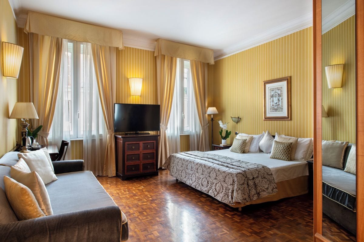 Una habitación de hotel con cama, sofá, televisor, cómoda y cortinas.