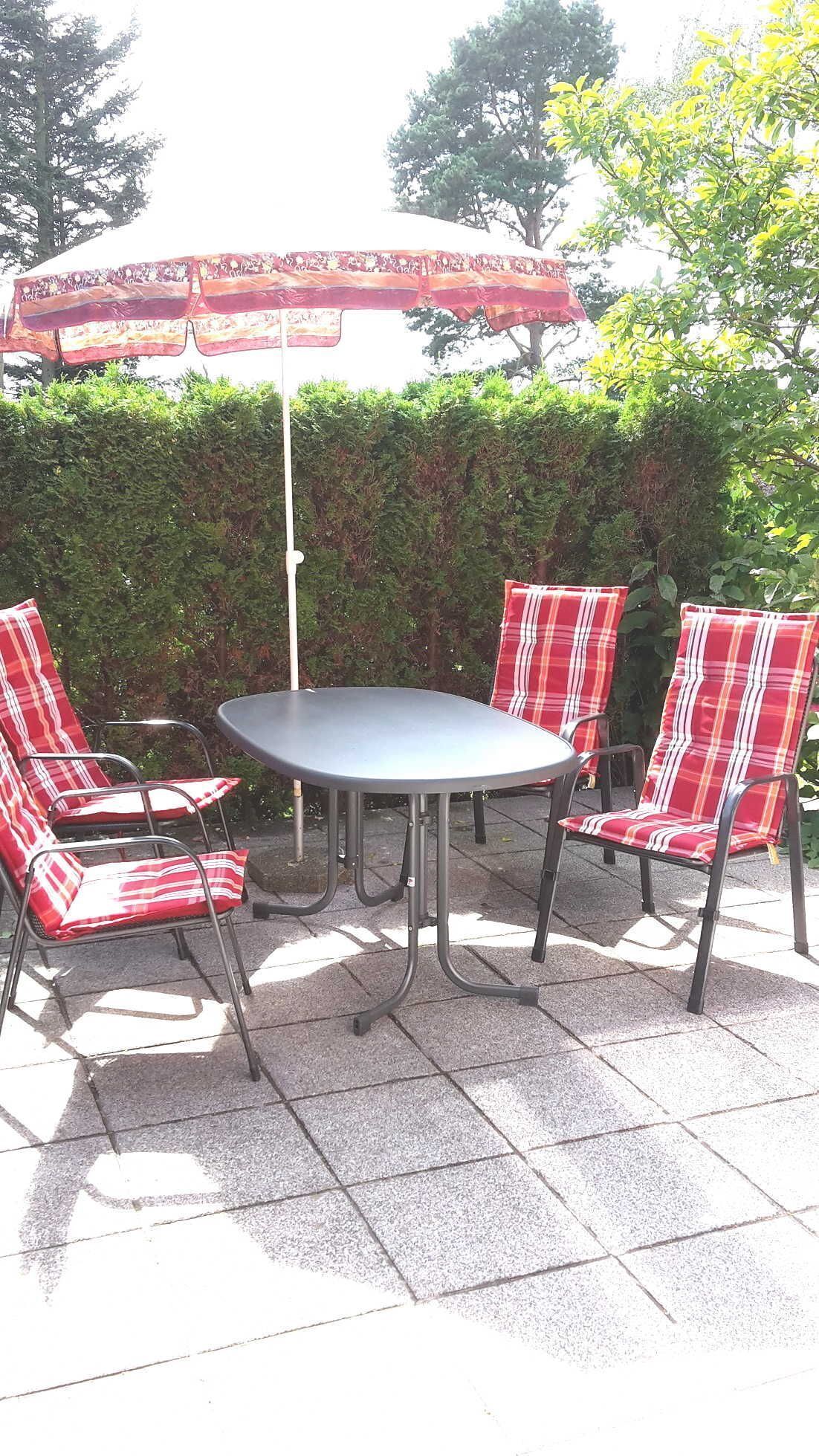 An outdoor patio with a round table and four plaid chairs, surrounded by a hedge and trees.