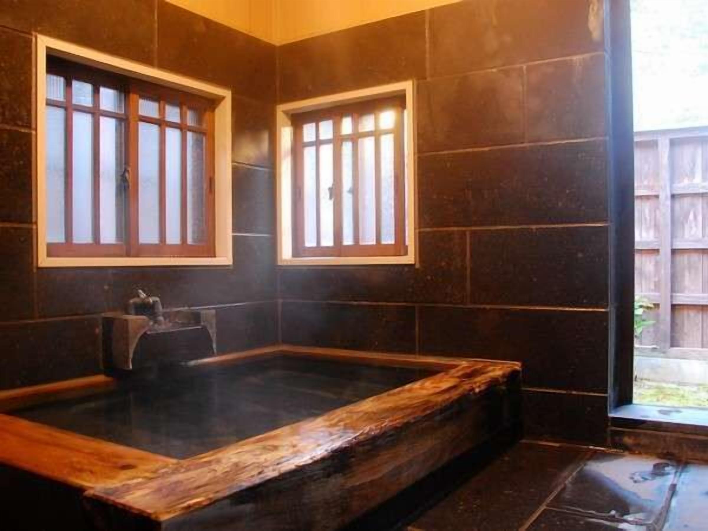 Habitación con una bañera de madera, paredes de azulejos oscuros y ventanas de madera.