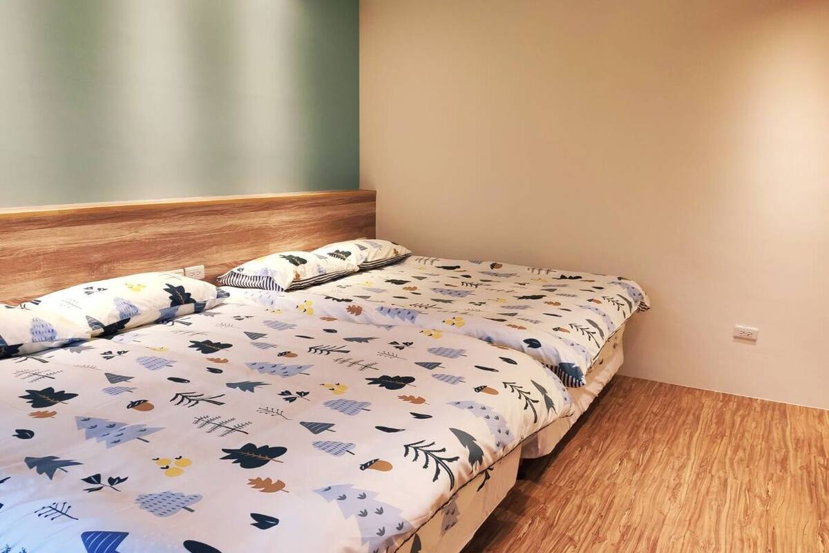 Una cama individual con colcha estampada, dos almohadas, cabecera de madera y pared lisa con interruptor de luz.