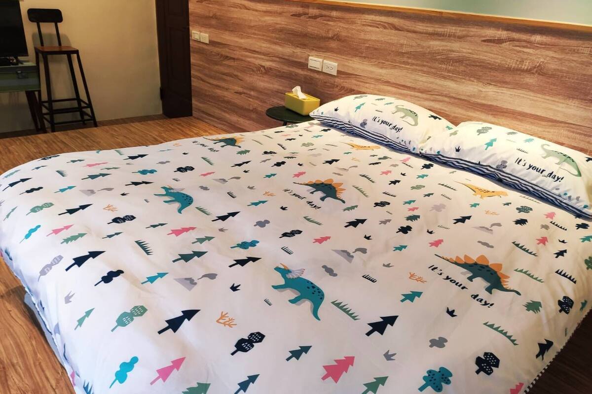 Una cama con una colcha temática de dinosaurios, una mesita de noche con una lámpara y una silla en una habitación de hotel.