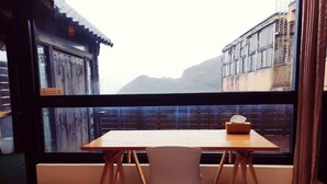 Una habitación con una mesa de madera y una silla, una ventana que ofrece vistas a edificios y montañas, y un cartel en la ventana.