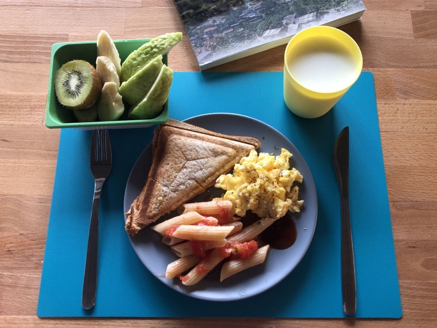 A plate with toast, scrambled eggs, and pasta, a green container with kiwi and green apples, and a yellow cup on a blue placemat.