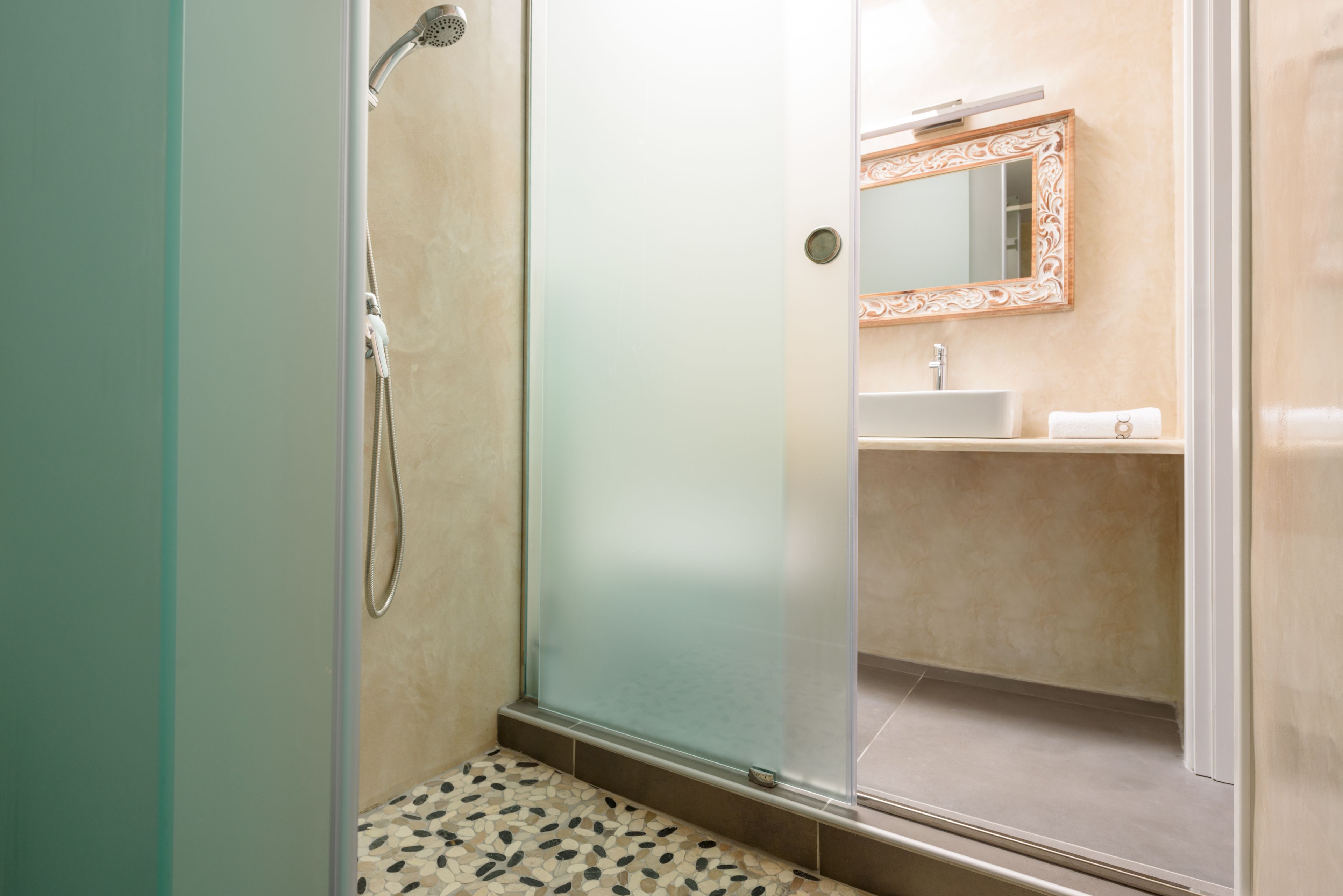 A glass-enclosed shower with a handheld showerhead, a tiled floor, and a bathroom with a sink and mirror.